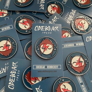 May include: A close-up of a stack of blue and white cards with a red and white enamel pin featuring a cartoon face with a lightning bolt. The text on the cards reads "ODEBEAR PRESS" and "ODEBEAR x ROBBIEXEFF" with the website address "www.odebearpress.com".