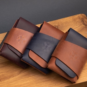 May include: Three leather wallets in different colors: burgundy, brown, and navy blue. Each wallet has a brown leather flap with a logo embossed on it.