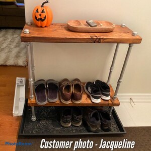 Peut inclure: Un porte-chaussures à deux niveaux en bois et en tuyaux métalliques. L'étagère supérieure comporte un plateau en bois et une petite citrouille orange. L'étagère inférieure comporte un plateau en métal noir. Le porte-chaussures est contre un mur blanc.