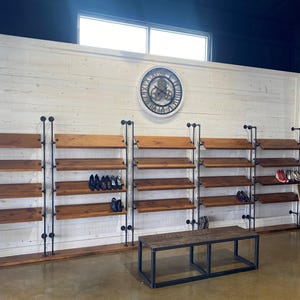 May include: A white wall with a large industrial clock and three sets of wooden shelves with black metal pipe supports. The shelves are filled with shoes. A black metal bench sits in front of the shelves.