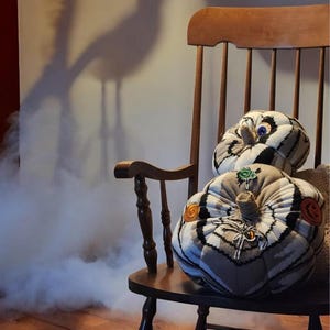 May include: Two fabric pumpkins with a black, white, and gray plaid pattern sit on a wooden rocking chair. The pumpkins feature decorative patches including a spider, a jack-o'-lantern, and a green emblem, with a single blue eye on the top pumpkin.