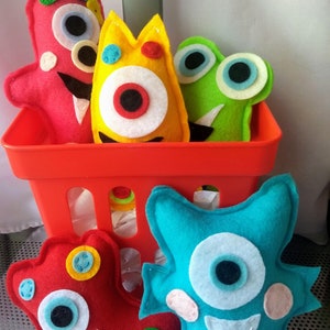May include: Five felt monster toys in a red plastic basket. The monsters are red, yellow, green, blue, and red with various colors and patterns. The monsters have one or two eyes and a variety of facial expressions.
