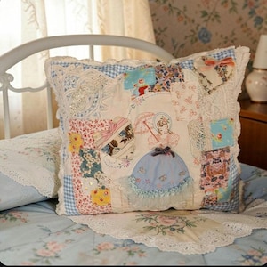 May include: A square decorative throw pillow featuring a patchwork design of various floral and gingham fabrics. The center displays an embroidered illustration of a person holding a parasol, accented with lace trim, ribbons, and fabric appliques.