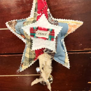 May include: A handmade star-shaped ornament with a patchwork design. The ornament features various fabric patterns in red, blue, and yellow, with a white center and the word "PEACE" on a plaid ribbon. Small silver bells hang from the bottom.