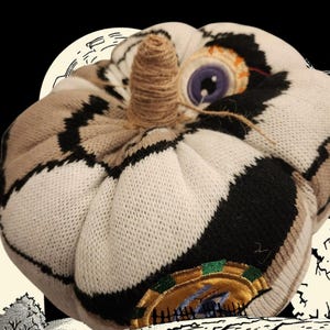 May include: A plush pumpkin crafted from knit fabric featuring a pattern of white, black, and tan segments. The top center features a stem wrapped in brown twine. An embroidered eye with a purple iris is visible on the upper right side.