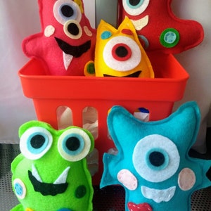 May include: Four felt monster toys in a red basket. The monsters are different colors and have different features, including eyes, teeth, and spots.