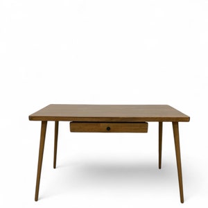 May include: A wooden desk with a single drawer and tapered legs. The desk is made of a light brown wood and has a natural finish. The drawer has a black knob. The desk is labeled "TRUMAKER".