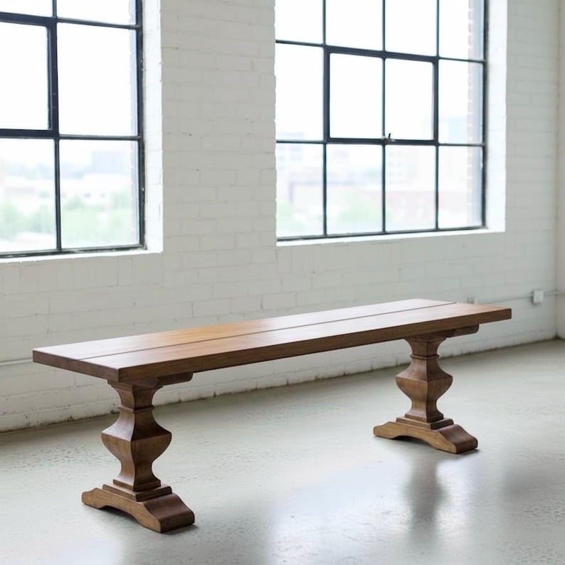 May include: A long, rectangular wooden bench featuring a smooth, flat seat surface supported by two ornate, pedestal-style legs. The bench is crafted from medium-toned brown wood with a visible grain, showcasing a sturdy, balanced design.