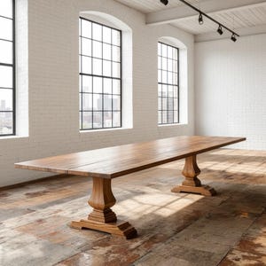 May include: A long rectangular wooden dining table featuring a smooth, natural brown grain surface. The table is supported by two substantial, carved pedestal bases with wide, tiered feet that provide a stable foundation for the tabletop.