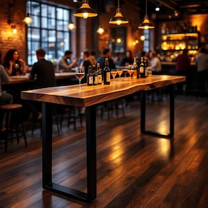 May include: A long rectangular bar table with a natural live edge wood top and black metal U-shaped legs. The table surface holds several glass bottles and stemmed glasses filled with amber liquid, positioned in a dimly lit indoor setting.