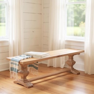 May include: A light-colored wooden bench with a rectangular top and ornate legs. A blue and white striped scarf and two books rest on the bench. The bench is in a room with white curtains and windows.