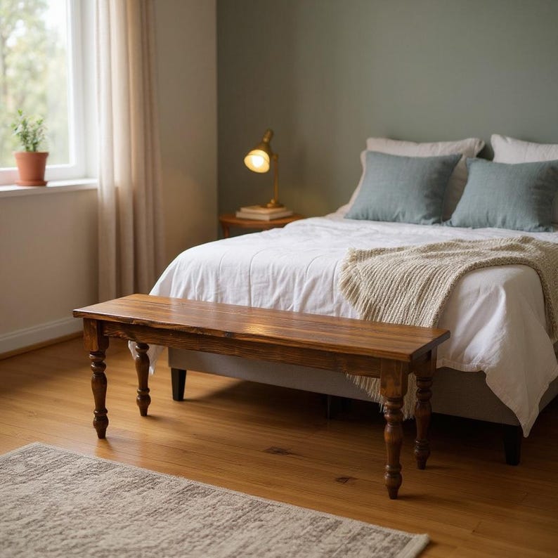May include: A wooden bench with a rich, brown finish. The bench has turned legs and is placed at the foot of a bed with white linens and blue pillows. A rug and a small table with a lamp are also visible.