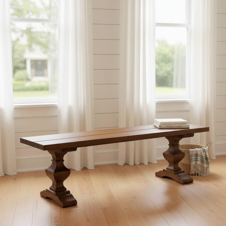 May include: A long, rectangular wooden bench with a smooth, dark brown finish. The bench features a flat seat supported by two thick, pedestal-style legs with a carved, curved silhouette. A small stack of books rests on the right side of the seat.