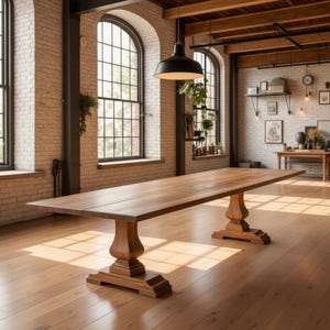 May include: A long rectangular wooden dining table with a smooth surface and two thick, carved pedestal bases. The table is crafted from medium-toned wood with a visible grain, featuring a sturdy construction that rests on wide, flat feet.