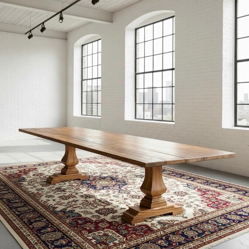 May include: A long rectangular wooden dining table with a smooth natural finish, featuring two thick pedestal bases with carved detailing. The table is positioned on a large, ornate area rug with a complex pattern in red, blue, and cream tones.