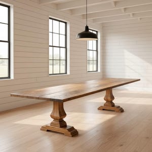 May include: A long rectangular wooden dining table featuring a smooth, natural brown grain surface and two sturdy pedestal bases with carved, tiered detailing. The table is positioned in a bright room with white horizontal wall paneling.