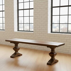 May include: A long, rectangular wooden bench featuring a smooth, dark brown finish. The bench is supported by two ornate, pedestal-style legs with a carved, bulbous design that tapers toward the base, providing a stable foundation for the seat.