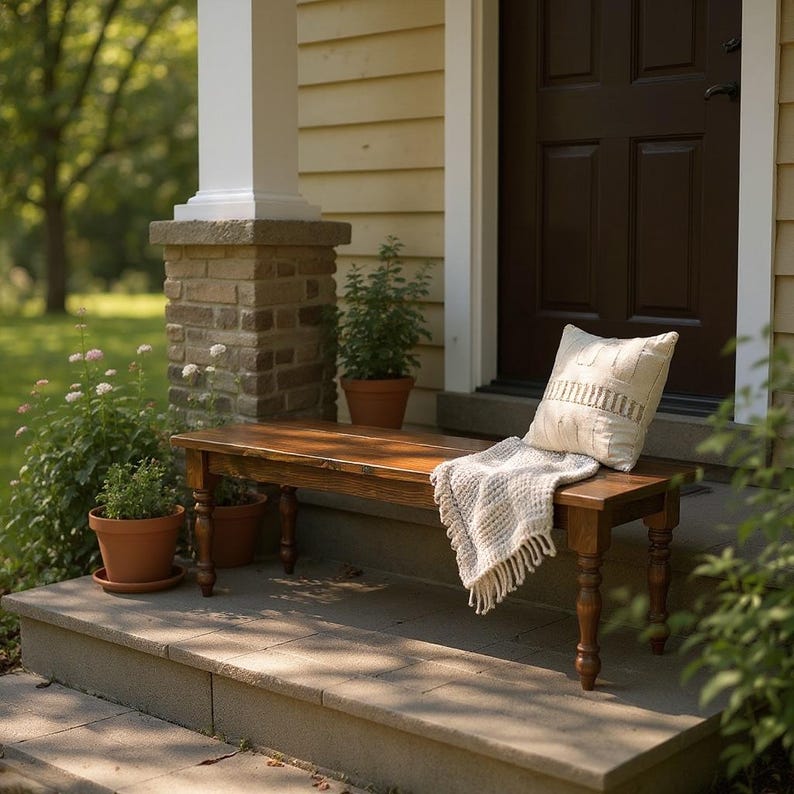 May include: A wooden bench with turned legs sits on a stone step. A cream-colored pillow and a fringed throw blanket rest on the bench. Potted plants and a brick and stone pillar are visible in the background.