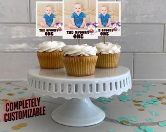 Könnte beinhalten: Drei Cupcakes mit weißer Zuckerglasur und einem Babyfoto auf einem Stiel, auf dem "The Spooky One" steht. Die Cupcakes stehen auf einem weißen Kuchenständer. Die Cupcakes stehen auf einer weißen Arbeitsplatte mit goldenen und türkisfarbenen Konfetti.