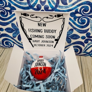 May include: White gift box with a fishing themed announcement that reads "New Fishing Buddy Coming Soon Baby Johnson October 2024". The box contains a red and white fishing bobber ornament with the text "Baby Johnson 2024".