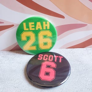 May include: Two round buttons. The top button is green with the name "LEAH" and the number "26" in yellow. The bottom button is black with the name "SCOTT" and the number "6" in pink.