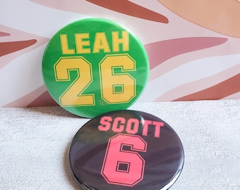 May include: Two round buttons. The top button is green with the name "LEAH" and the number "26" in yellow. The bottom button is black with the name "SCOTT" and the number "6" in pink.