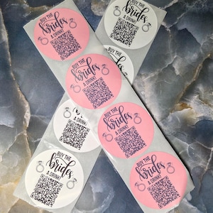 May include: A collection of round stickers, some pink and some white, with the text "BUY THE BRIDES A DRINK!" in a script font. Each sticker features a QR code and a ring graphic.