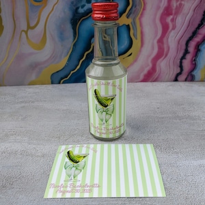 May include: A small glass bottle with a red cap, featuring a label with a pickle martini design and the text "Nicole's Bachelorette August 30, 2025." A matching card is next to the bottle. The label has green and white stripes.
