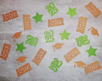 May include: A selection of glittery orange and green graduation-themed cutouts scattered on a light grey surface. The cutouts include the name "Asher", the year "2025", stars, and graduation caps. The items are likely for a graduation celebration.