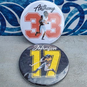 May include: Two round buttons with sports-themed designs. One button is white with the name "Anthony" and the number "33" in orange, featuring a football player. The other is black with "Johnson" and "17" in yellow, showing a baseball player.