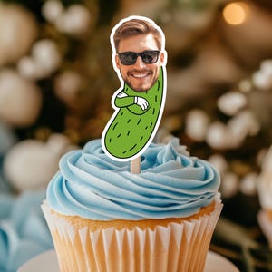 May include: A cupcake with light blue frosting and a decorative topper. The topper features a cartoon image of a man's face wearing sunglasses, with his face on a green pickle.