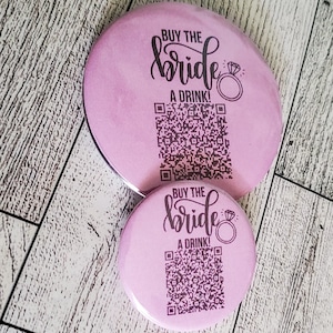 May include: Two pink round buttons with the text "Buy the bride a drink!" and a diamond ring icon. Each button has a black and white QR code.