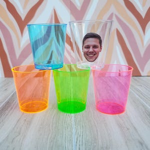 May include: Four clear plastic cups, one with a face cutout, in different colors: orange, green, pink, and blue. The cups are arranged in a row on a wooden surface.
