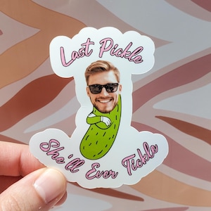 May include: A white sticker featuring a smiling man's face wearing sunglasses, integrated into a green pickle design. The sticker has pink text that reads "Last Pickle She'll Ever Tickle."