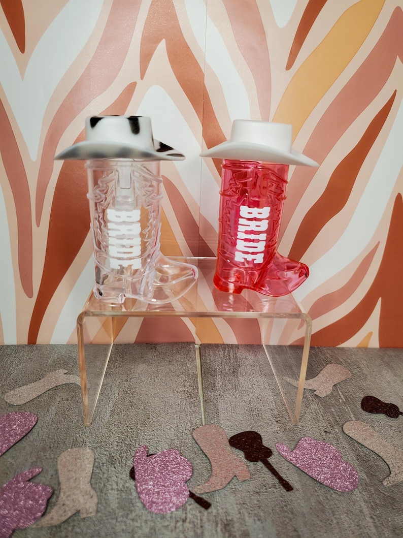 K&ouml;nnte beinhalten: Zwei durchsichtige Cowboy-Stiefel-f&ouml;rmige Becher aus Plastik mit wei&szlig;en Cowboyh&uuml;ten oben drauf. Die Becher sind mit dem Wort "BRIDE" in wei&szlig;en Buchstaben verziert. Die Becher stehen auf einem durchsichtigen Acrylregal.