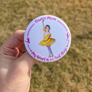 May include: A round button with the text "Dance Mom" and "My heart is on that stage" in pink. The button features a photo of a young girl in a yellow tutu. Arrows and ballerina silhouettes decorate the border.