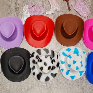 K&ouml;nnte beinhalten: Neun bunte Cowboyh&uuml;te in verschiedenen Mustern und Farben, darunter lila, rot, braun, rosa, wei&szlig;, schwarz und ein schwarz-wei&szlig;es Kuhmuster.