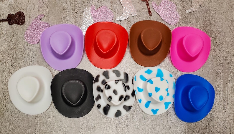 K&ouml;nnte beinhalten: Neun bunte Cowboyh&uuml;te in verschiedenen Mustern und Farben, darunter lila, rot, braun, rosa, wei&szlig;, schwarz und ein schwarz-wei&szlig;es Kuhmuster.