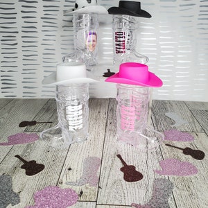 K&ouml;nnte beinhalten: Vier durchsichtige Cowboy-Stiefel-f&ouml;rmige Becher aus Plastik mit verschiedenen farbigen Cowboyh&uuml;ten oben drauf. Die H&uuml;te sind rosa, wei&szlig;, schwarz und wei&szlig; mit schwarzen Punkten. Die Becher stehen auf einer Holzoberfl&auml;che mit Glitzerkonfetti.