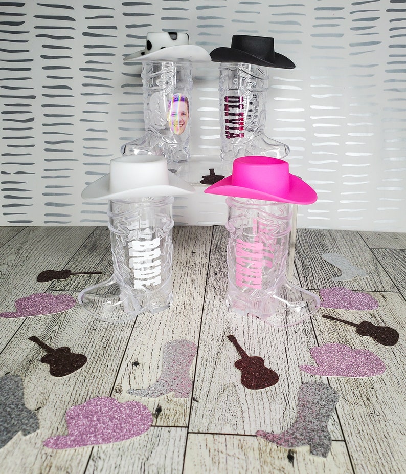 K&ouml;nnte beinhalten: Vier durchsichtige Cowboy-Stiefel-f&ouml;rmige Becher aus Plastik mit verschiedenen farbigen Cowboyh&uuml;ten oben drauf. Die H&uuml;te sind rosa, wei&szlig;, schwarz und wei&szlig; mit schwarzen Punkten. Die Becher stehen auf einer Holzoberfl&auml;che mit Glitzerkonfetti.