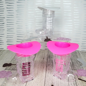 K&ouml;nnte beinhalten: Zwei durchsichtige Cowboy-Stiefel-Schnapsgl&auml;ser aus Plastik mit rosa Cowboyh&uuml;ten. Die Gl&auml;ser haben den Schriftzug "Bride" aufgedruckt.