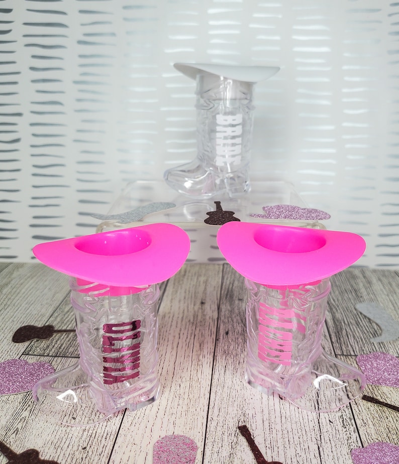 K&ouml;nnte beinhalten: Zwei durchsichtige Cowboy-Stiefel-Schnapsgl&auml;ser aus Plastik mit rosa Cowboyh&uuml;ten. Die Gl&auml;ser haben den Schriftzug "Bride" aufgedruckt.
