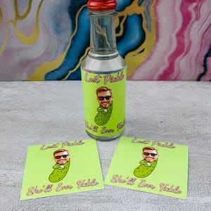 May include: A clear glass bottle with a red cap and a green label featuring a cartoon pickle with a face and sunglasses. The label reads "Last Pickle" and "She'll Ever Tickle." Two matching coasters are also present.