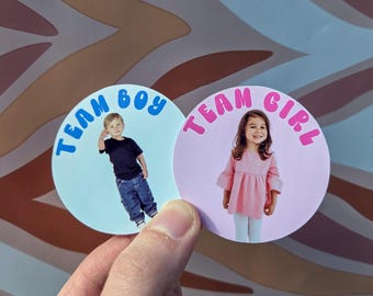 Könnte beinhalten: Zwei runde Aufkleber werden in einer Hand gehalten. Einer zeigt einen Jungen und die Worte "TEAM BOY" in Blau. Der andere zeigt ein Mädchen und die Worte "TEAM GIRL" in Pink. Ideal für eine Geschlechtsenthüllung.
