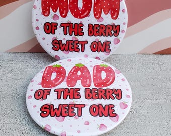 May include: Two round white buttons with red text and strawberry graphics. One button says "MOM of the berry sweet one" and the other says "DAD of the berry sweet one". The buttons have a pink strawberry pattern.