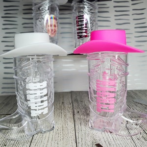 K&ouml;nnte beinhalten: Zwei durchsichtige Cowboy-Stiefel-f&ouml;rmige Becher aus Plastik mit Deckeln. Ein Becher hat einen wei&szlig;en Cowboyhut-Deckel und das Wort "Bride" auf dem Stiefel geschrieben. Der andere Becher hat einen rosafarbenen Cowboyhut-Deckel und das Wort "Maid of Honor" auf dem Stiefel geschrieben.