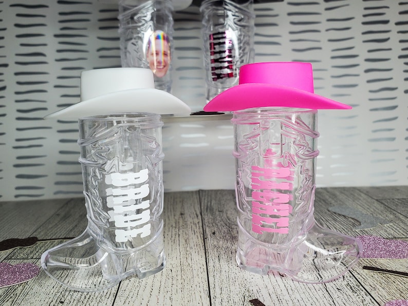 K&ouml;nnte beinhalten: Zwei durchsichtige Cowboy-Stiefel-f&ouml;rmige Becher aus Plastik mit Deckeln. Ein Becher hat einen wei&szlig;en Cowboyhut-Deckel und das Wort "Bride" auf dem Stiefel geschrieben. Der andere Becher hat einen rosafarbenen Cowboyhut-Deckel und das Wort "Maid of Honor" auf dem Stiefel geschrieben.