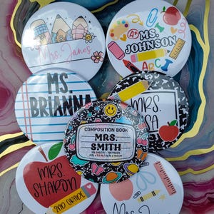May include: A collection of round teacher-themed buttons. Designs include pencils, apples, and school supplies. Each button features a teacher's name, such as "Mrs. Smith" or "Ms. Johnson." The buttons are white with colorful graphics and text.