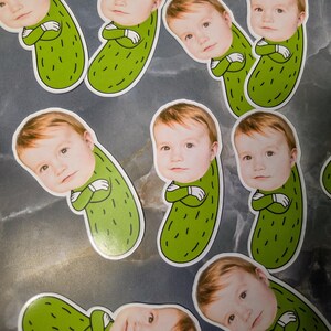 以下が含まれることがあります： 緑色のピクルスの体に赤ちゃんの顔が描かれたステッカーのコレクション。ステッカーには白い縁取りがあり、赤ちゃんの腕は組まれています。背景はまだらのグレーとシルバーの表面です。