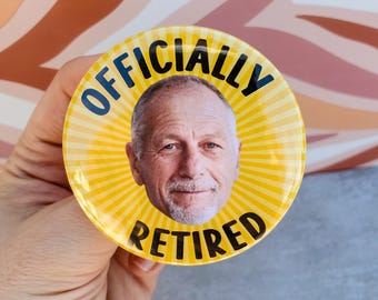 Puede incluir: Un botón redondo amarillo con el texto "OFFICIALLY RETIRED" en negro y la cara de un hombre sonriente en el centro. El botón tiene un diseño de rayos y está sostenido por una mano.